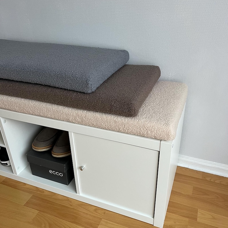 May include: A white storage bench with three plush, textured cushions in gray, brown, and beige. The bench has open cubbies and a cabinet with a silver knob. A pair of shoes and a black shoe box are visible inside the bench.