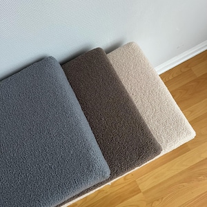 May include: Three rectangular rugs in a row, each with a textured, plush surface. The rugs are in shades of gray, brown, and beige. The rugs are placed on a wooden floor, with a white wall in the background.