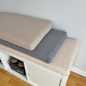 May include: Three rectangular, plush cushions in beige, gray, and cream colors are stacked on a white cabinet. The cushions have a soft, textured appearance. A pair of brown shoes is visible inside the cabinet.