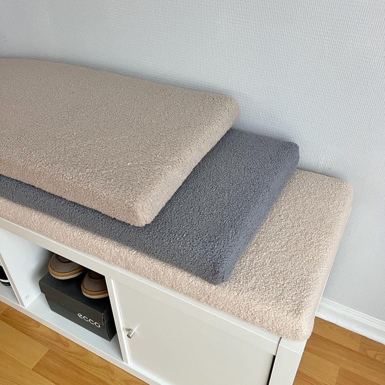 May include: Three rectangular, plush cushions in beige, gray, and cream colors are stacked on a white cabinet. The cushions have a soft, textured appearance. A pair of brown shoes is visible inside the cabinet.