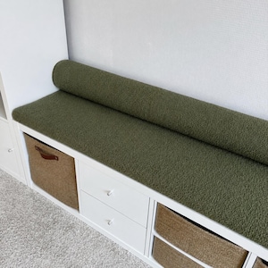 May include: A white storage unit with an olive green textured bench seat. The bench has a rolled-up section at the back. Woven baskets and white drawers are visible below the bench.