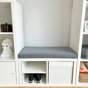 May include: A white storage unit with a built-in bench featuring a gray cushion. Below the bench are open shelves, a white storage box, and a pair of black and white sneakers. Decorative items are on the shelves.
