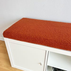 May include: A white storage cabinet with a burnt orange, textured cushion on top. The cabinet has a door with a small, silver handle and an open shelf. The cushion appears to be made of a soft, plush material.