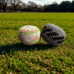 Puede incluir: Dos pelotas de malabares tejidas a ganchillo de forma redonda. Una pelota es de color gris claro con un patrón repetitivo de puntos rosa neón y verde neón. La segunda es de color gris oscuro sólido con un patrón negro texturizado tejido.