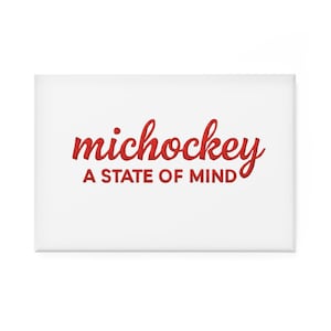 Puede incluir: Un imán rectangular blanco con las palabras "michockey" en rojo en escritura cursiva sobre la frase "A STATE OF MIND" en letras rojas. El diseño es simple y limpio.