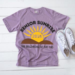 May include: A lilac-colored t-shirt with the words "SENIOR SUNRISE 2026" in black and yellow, featuring a sun graphic. Below the graphic is the phrase "THE BEGINNING OF THE END." White sneakers are placed next to the shirt.