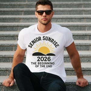 May include: White t-shirt with the text "SENIOR SUNRISE 2026 THE BEGINNING OF THE END" in black and yellow. The design features a yellow sun rising over a black mountain range. The shirt is worn by a person.