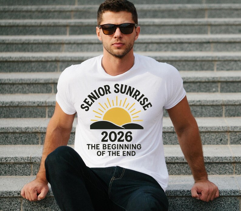 May include: White t-shirt with the text "SENIOR SUNRISE 2026 THE BEGINNING OF THE END" in black and yellow. The design features a yellow sun rising over a black mountain range. The shirt is worn by a person.