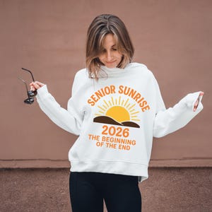 May include: White hooded sweatshirt with the text "SENIOR SUNRISE 2026 THE BEGINNING OF THE END" in orange and yellow. The design features a sun rising over a brown landscape. The sweatshirt is worn by a person.