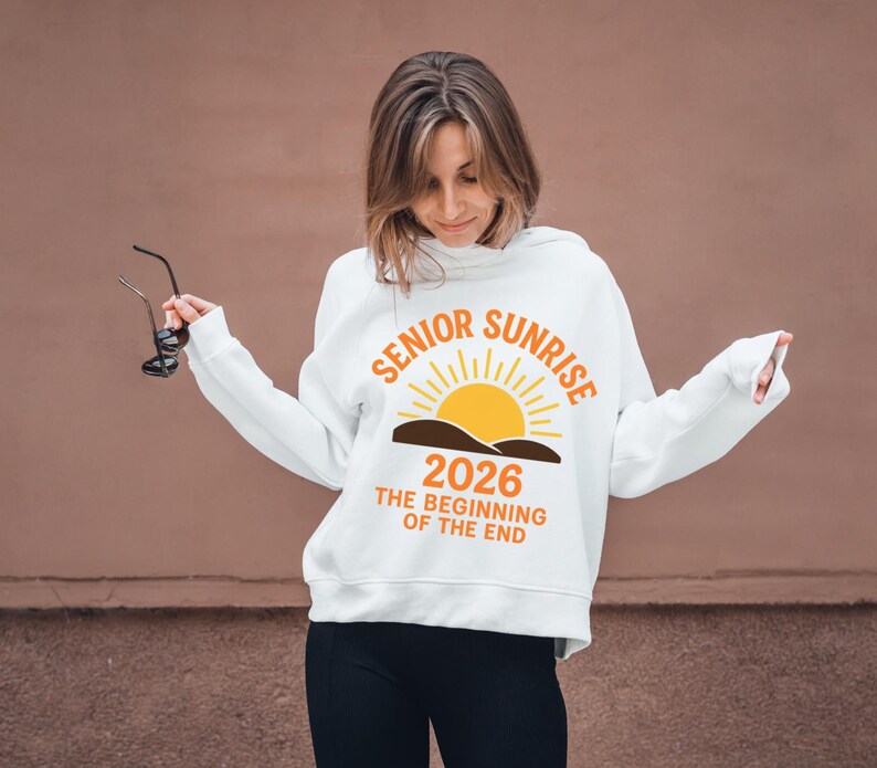 May include: White hooded sweatshirt with the text "SENIOR SUNRISE 2026 THE BEGINNING OF THE END" in orange and yellow. The design features a sun rising over a brown landscape. The sweatshirt is worn by a person.