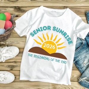 May include: White t-shirt with the text "SENIOR SUNRISE 2026" in teal and yellow. The design includes a yellow sun, brown hills, and the phrase "THE BEGINNING OF THE END" in teal. The shirt is on a wooden surface.