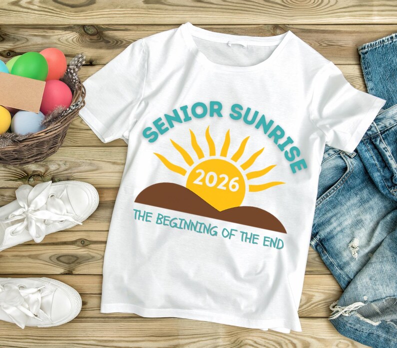 May include: White t-shirt with the text "SENIOR SUNRISE 2026" in teal and yellow. The design includes a yellow sun, brown hills, and the phrase "THE BEGINNING OF THE END" in teal. The shirt is on a wooden surface.