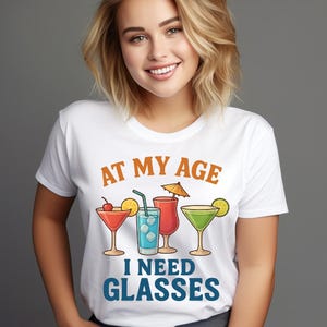 Può includere: T-shirt bianco con la scritta "AT MY AGE I NEED GLASSES" in arancione e blu. La maglietta presenta illustrazioni di quattro diversi cocktail con guarnizioni, tra cui una ciliegia, una fetta di limone e un ombrellino.