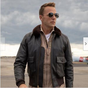 Könnte beinhalten: Eine braune Lederbomberjacke mit einem Pelzkragen und Reißverschluss. Die Jacke hat zwei große Taschen auf der Vorderseite und eine Reißverschlusstasche am linken Ärmel. Das Model trägt die Jacke mit einem hellbraunen Hemd darunter.