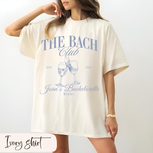 Puede incluir: Camiseta de color marfil con el texto "THE BACH Club" en azul claro, junto con las palabras "Joan's Bachelorette" y "Miami". Se representan dos copas de vino, con el año "2025" y la palabra "EST." también incluida.