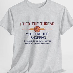 May include: A light gray t-shirt with the text "I TIED THE THREAD, YOU FUND THE SHOPPING" in maroon and navy blue. The shirt also has a graphic of a Rakhi. The text "THE RAKHI MAY BE SMALL, BUT THE EMOTIONAL DAMAGE IS MASSIVE" is also on the shirt.