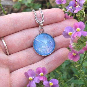 May include: A silver-toned necklace with a round pendant featuring a blue and glittery design. The pendant hangs from a twisted rope chain. The necklace is displayed with purple flowers in the background.