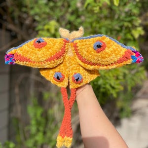 May include: A handmade, plush moth toy with yellow, orange, blue, and pink accents. The moth has large, textured wings with blue and orange details, and a pair of orange legs. The moth's body is yellow with a small, tan head.