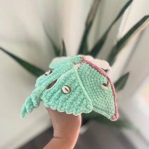 May include: A handmade crochet moth in shades of mint green, pink, and white. The moth has a textured, plush appearance with button accents. The wings are detailed with pink edging. The moth is held in front of a blurred background of green leaves.