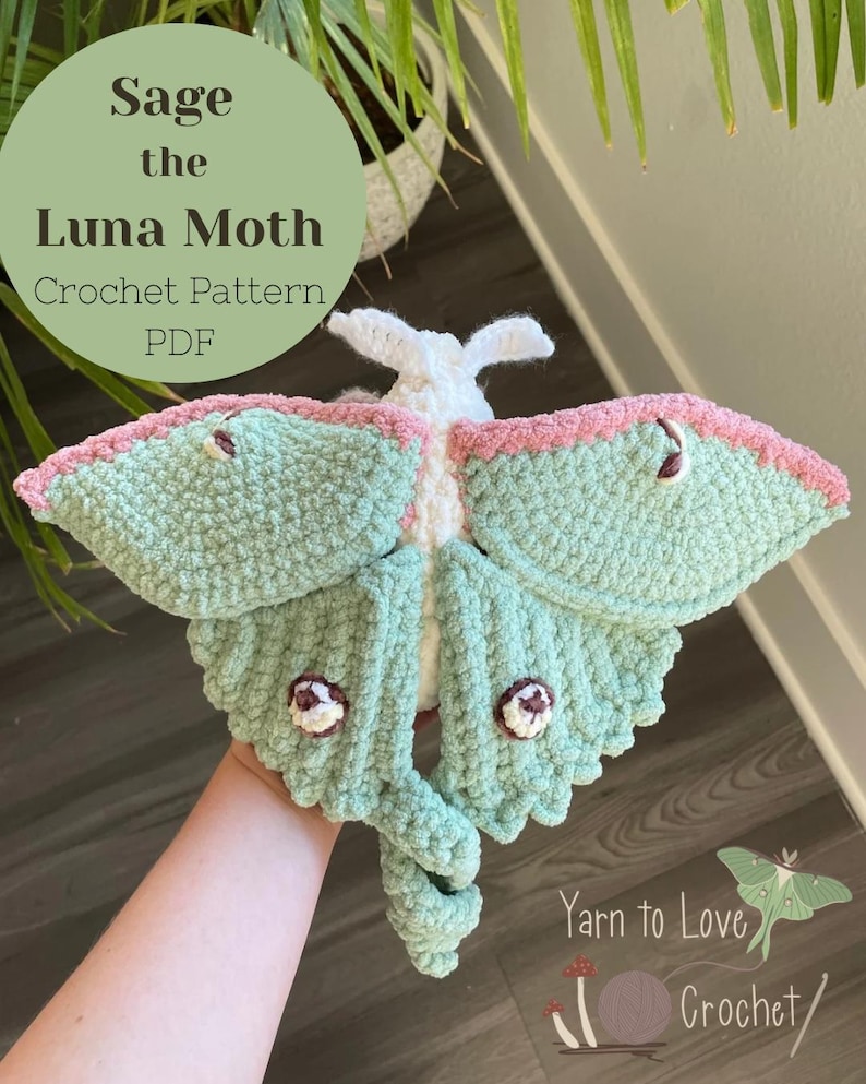 May include: A crocheted Luna moth in shades of sage green, pink, and white. The moth has detailed wings and a textured body. The image includes the text "Sage the Luna Moth Crochet Pattern PDF".
