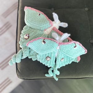 May include: Two handmade, plush Luna moths in soft teal, pink, and white. The moths have detailed wings and antennae, with small, button-like details. The plush toys are resting on a dark green surface.