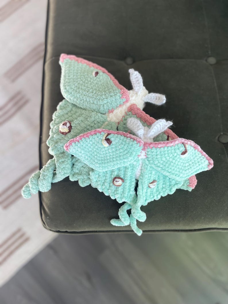 May include: Two handmade, plush Luna moths in soft teal, pink, and white. The moths have detailed wings and antennae, with small, button-like details. The plush toys are resting on a dark green surface.