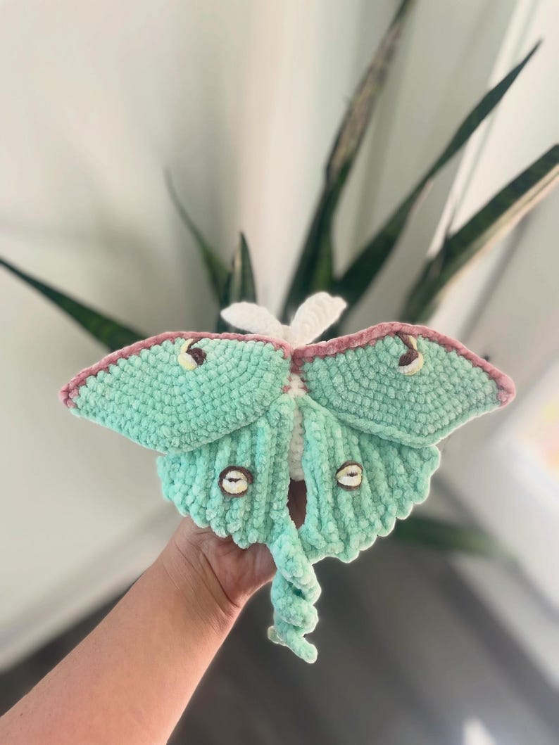 May include: A handmade crochet moth in shades of mint green, pink, and white. The moth has detailed wings, brown eye-like markings, and a curled tail. The plush toy is soft and textured, perfect for home decor or as a unique gift.