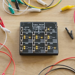 Puede incluir: Una placa de circuito Logic Gates negra con varios componentes electrónicos, incluidos cables y pinzas de cocodrilo. La placa presenta puertas lógicas etiquetadas como AND, OR y NOT, con luces iluminadas. Los cables en rojo, amarillo, verde y blanco están dispersos.