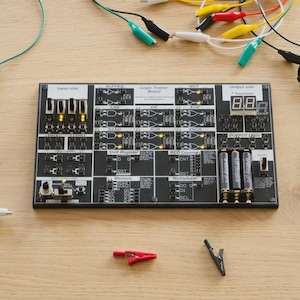 May include: A black Logic Trainer Board with electronic components, LEDs, switches, and a digital display. Various coloured wires with alligator clips are connected to the board. The board is on a light brown wooden surface.