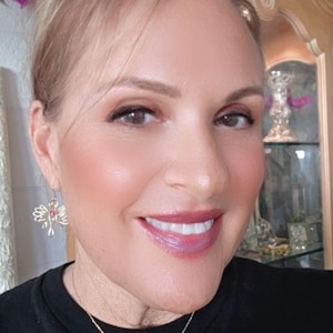 May include: A woman wearing gold angel-shaped earrings and a matching pendant necklace with a pink gemstone. The woman is wearing a black top and has a floral hair accessory with purple roses.