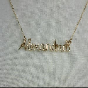 Pode incluir: Um colar de ouro com o nome "Alexandra" escrito em cursivo. O colar est&aacute; sobre um fundo branco.