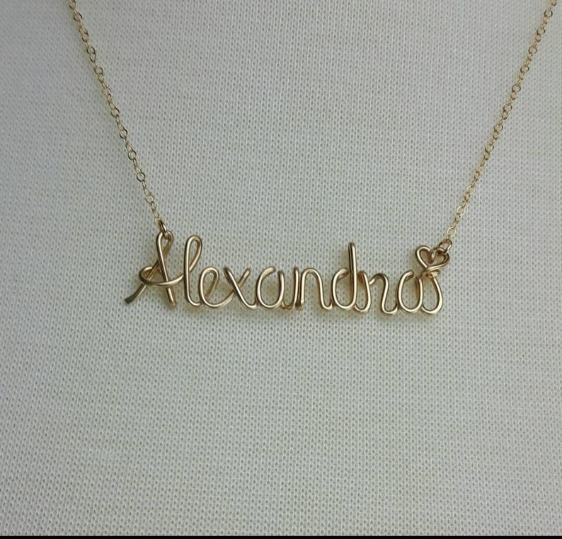 Pode incluir: Um colar de ouro com o nome "Alexandra" escrito em cursivo. O colar est&aacute; sobre um fundo branco.
