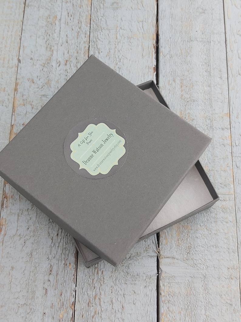 May include: A gray square box with a white label that says "A Gift for You from...  Joanne Watson Jewelry" and a website address. The box is open and the inside is lined with white paper.