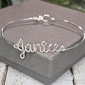 May include: A silver-toned wire bracelet with the name "Janice" in cursive script. The bracelet is displayed on a gray box, showcasing the delicate craftsmanship and personalized design. The bracelet's clasp is visible.