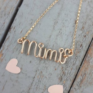 May include: Gold necklace with a wire-wrapped 'Mimi' pendant. The necklace is on a wooden surface with pink hearts.