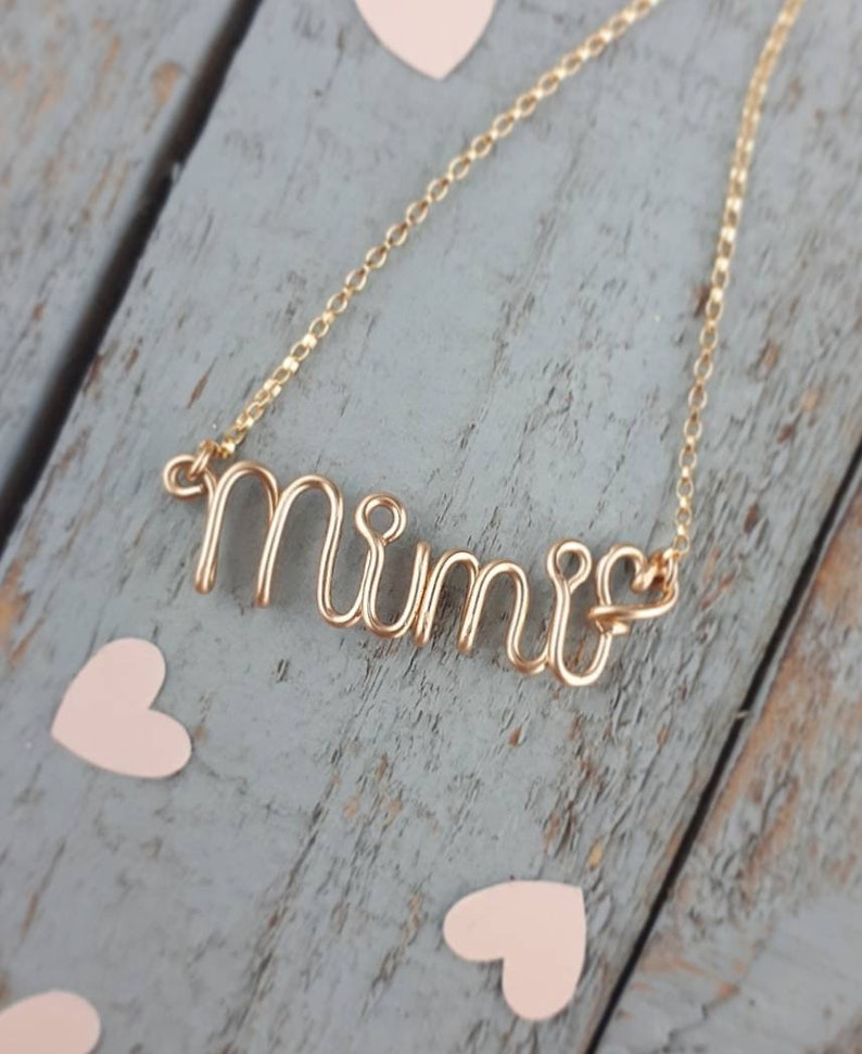 May include: Gold necklace with a wire-wrapped 'Mimi' pendant. The necklace is on a wooden surface with pink hearts.