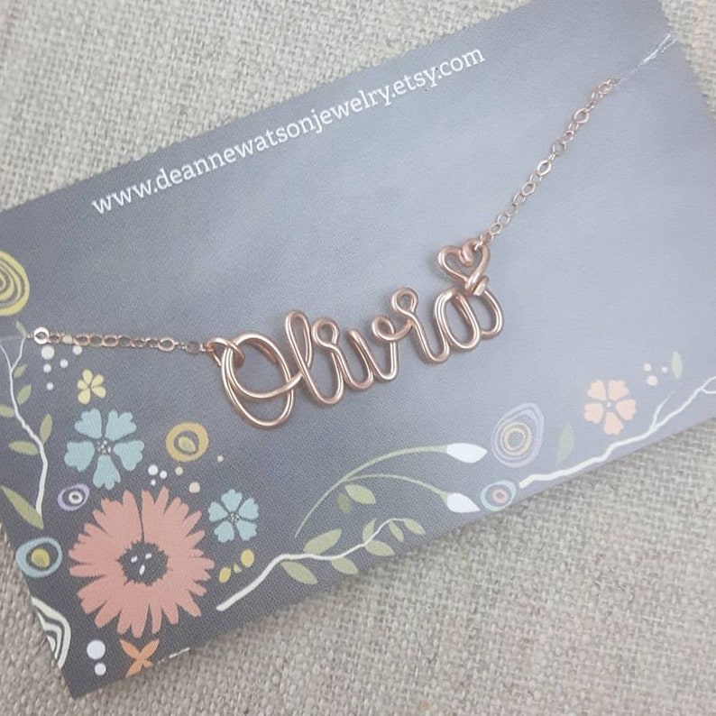 Pode incluir: Um colar de ouro rosa com o nome "Olivia" escrito em arame. Um pequeno pingente em forma de cora&ccedil;&atilde;o est&aacute; preso ao colar.