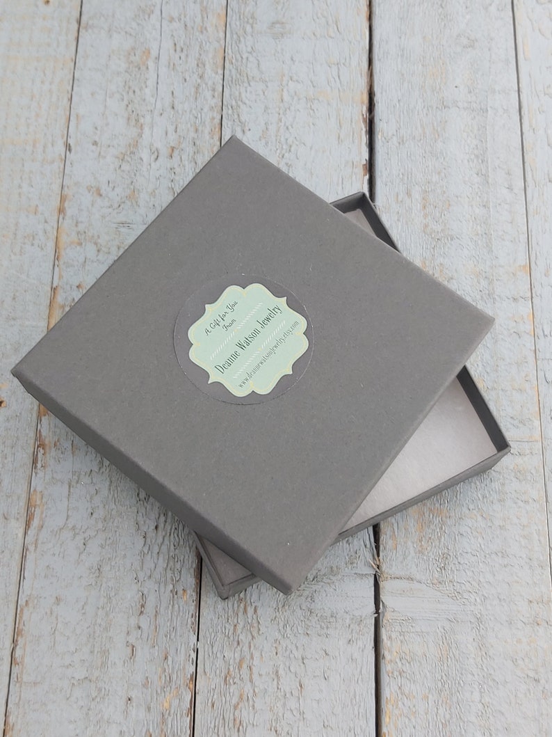 May include: A grey gift box, slightly ajar, revealing a light pink interior. The box rests on a weathered, light blue wooden surface. A sticker on the lid reads "A Gift for You From Deanne Watson Jewelry".
