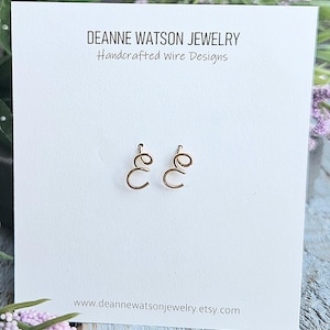 May include: A pair of gold-colored wire earrings in the shape of the letter "e" on a white card. The card has the text "DEANNE WATSON JEWELRY Handcrafted Wire Designs" and the website address.