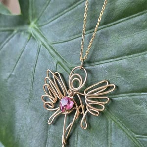 May include: A gold-coloured wire angel pendant necklace with a pink gemstone centre. The angel has wire wings and a delicate chain. The necklace is displayed on a large green leaf.
