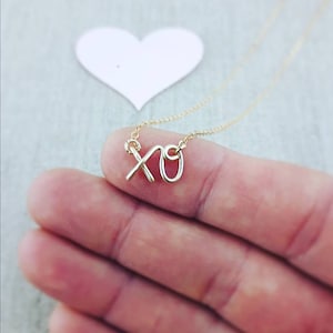 May include: A gold necklace with a delicate XO charm. The charm is made of thin wire and is shaped like the letters XO. The necklace is on a chain and is being held by a hand.