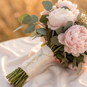 May include: A bouquet of pale pink peonies and green eucalyptus leaves with stems wrapped in cream-colored ribbon. A delicate gold-toned chain with a small blue bead and a charm featuring the cursive text Mrs. Rogers is attached to the ribbon.