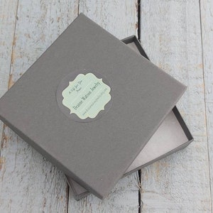 Może przedstawiać: Szare kwadratowe pudełko z białą etykietą, na kt&oacute;rej widnieje napis "Dreamie Watson Jewelry" oraz "A Gift for You from Dreamie". Etykieta ma ozdobną ramkę.