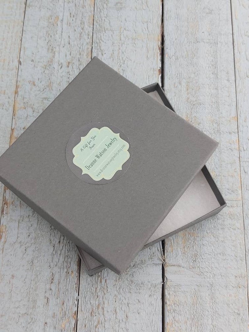 Może przedstawiać: Szare kwadratowe pudełko z białą etykietą, na kt&oacute;rej widnieje napis "Dreamie Watson Jewelry" oraz "A Gift for You from Dreamie". Etykieta ma ozdobną ramkę.