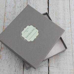 Pode incluir: Uma caixa quadrada cinza com um adesivo branco que diz "Dreamie Watson Jewelry" e "A Gift for You from Dreamie Watson Jewelry" com um endere&ccedil;o de site. A caixa est&aacute; aberta e o interior &eacute; forrado com papel branco.