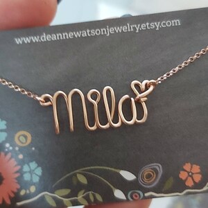 May include: A rose gold necklace with the name "Mila" spelled out in wire, with a small heart charm at the end of the name. The necklace is on a black card with a floral pattern and the website address "www.deannenewatsonjewelry.etsy.com".