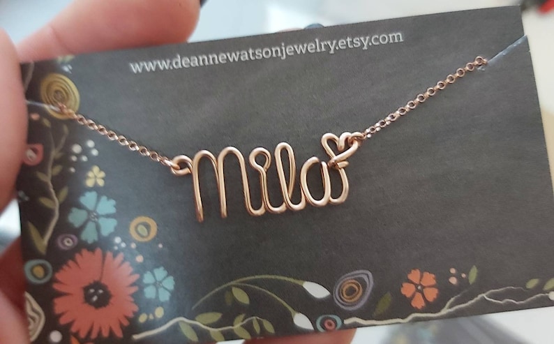 May include: A rose gold necklace with the name "Mila" spelled out in wire, with a small heart charm at the end of the name. The necklace is on a black card with a floral pattern and the website address "www.deannenewatsonjewelry.etsy.com".