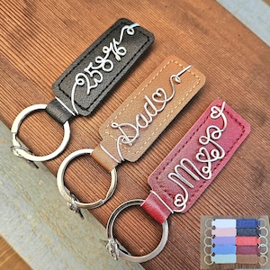 May include: Three personalized keychains with silver wire lettering on leather tags. The keychains are black, brown, and burgundy. The text on the keychains reads "258%", "Dad", and "Mrs". A small display of additional keychains is in the bottom right corner.