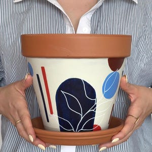 Könnte beinhalten: Ein terrakottafarbener Keramik-Blumentopf mit passender Untertasse. Der Topf ist mit abstrakten Formen in Marineblau, Rot und Hellblau auf einem cremefarbenen Hintergrund bemalt. Eine Person mit gestreiftem Hemd hält den Topf.