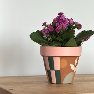 May include: A decorative plant pot in terracotta, featuring a pink rim and geometric patterns in green, pink, and white. It holds a flowering plant with purple flowers and green leaves. The pot is placed on a wooden surface.